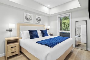 A modern bedroom with a large bed, two bedside tables, a nightstand, and a window with a view of greenery.