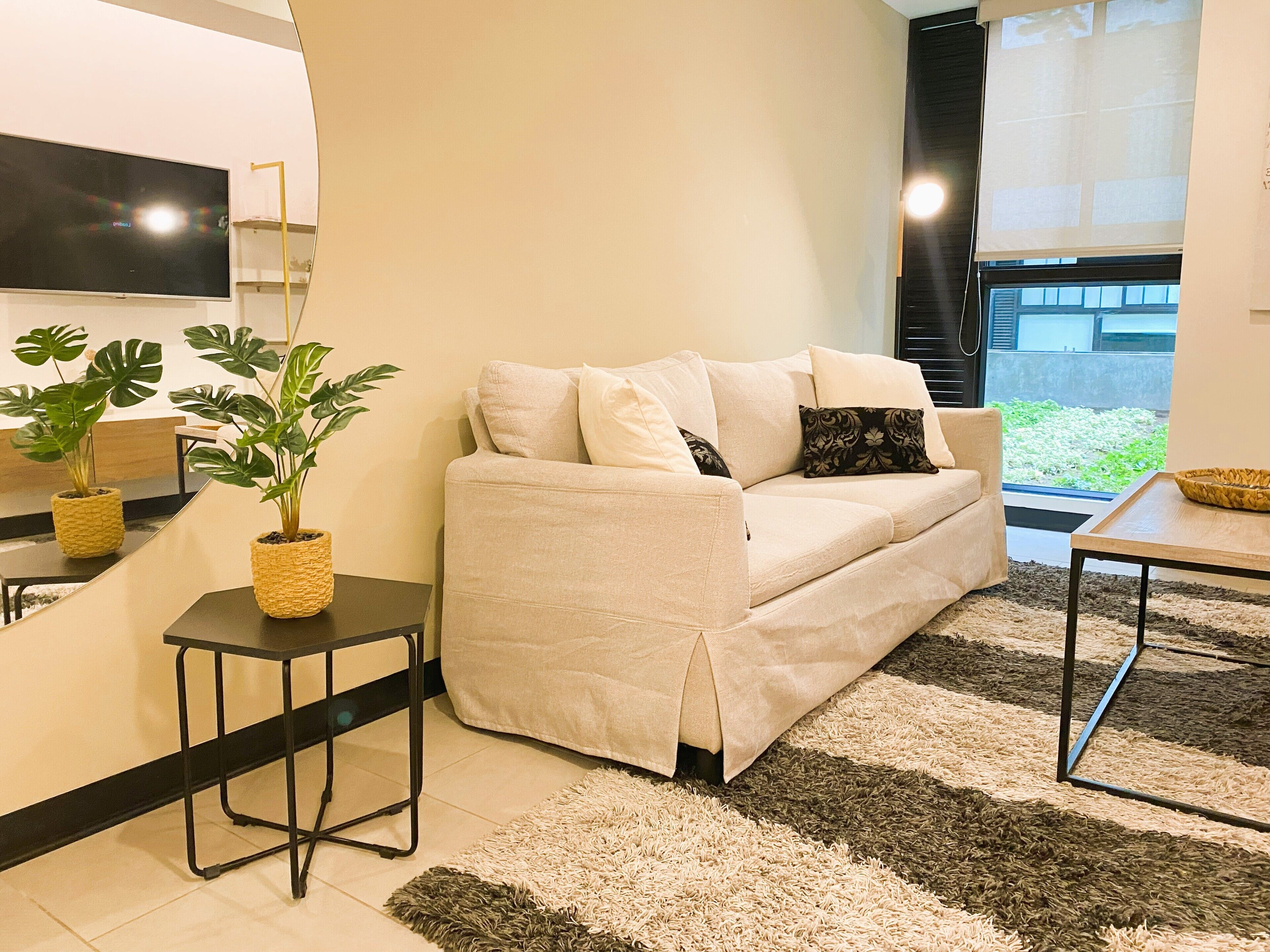 A modern living room with a beige sofa, two potted plants, a small black table, and a large window with a view of greenery outside.