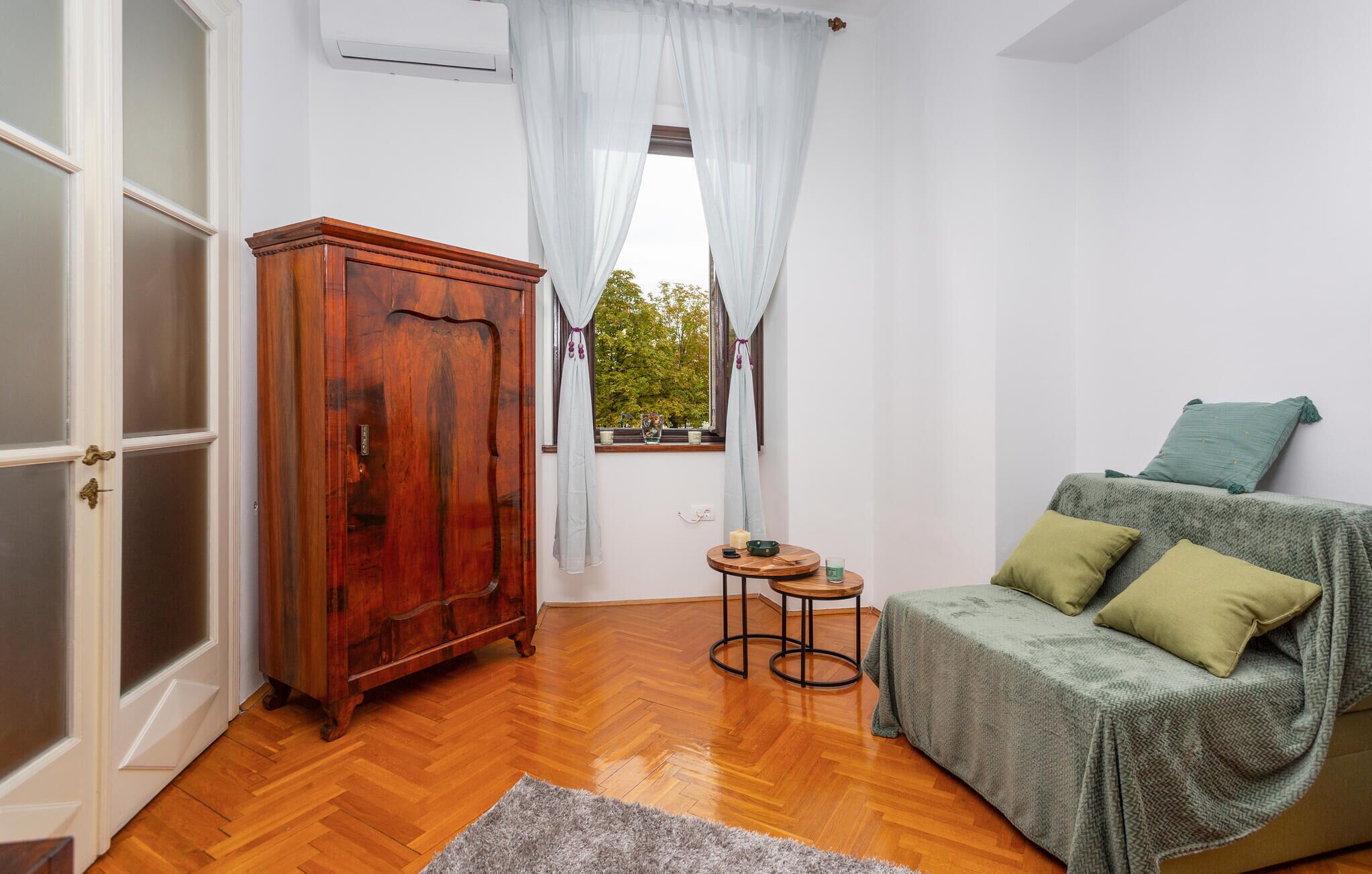 A room with a wooden wardrobe, a small round table, a green sofa with cushions, and a window with light curtains.