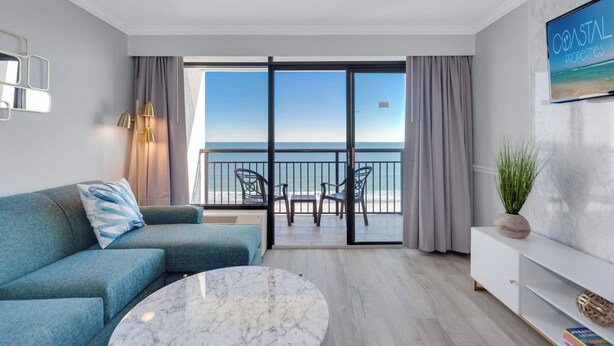 A modern hotel room with a balcony, a sofa, a round table, and a TV displaying 'Coastal Properties'.