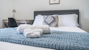 Una cama bien hecha con una manta azul con textura y toallas blancas enrolladas, un cabecero gris y un cuadro enmarcado en la pared.