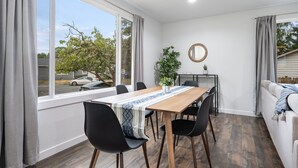 Un comedor con mesa de madera, sillas negras, una planta en maceta y vistas a la calle a través de amplias ventanas.