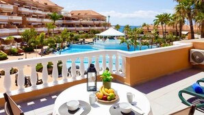 A round table with a white tablecloth, surrounded by chairs, overlooking a swimming pool with a view of a hotel building and palm trees. - Home2Book Modern Seaview Apt & Pool, Las Américas (Playa de la Américas)