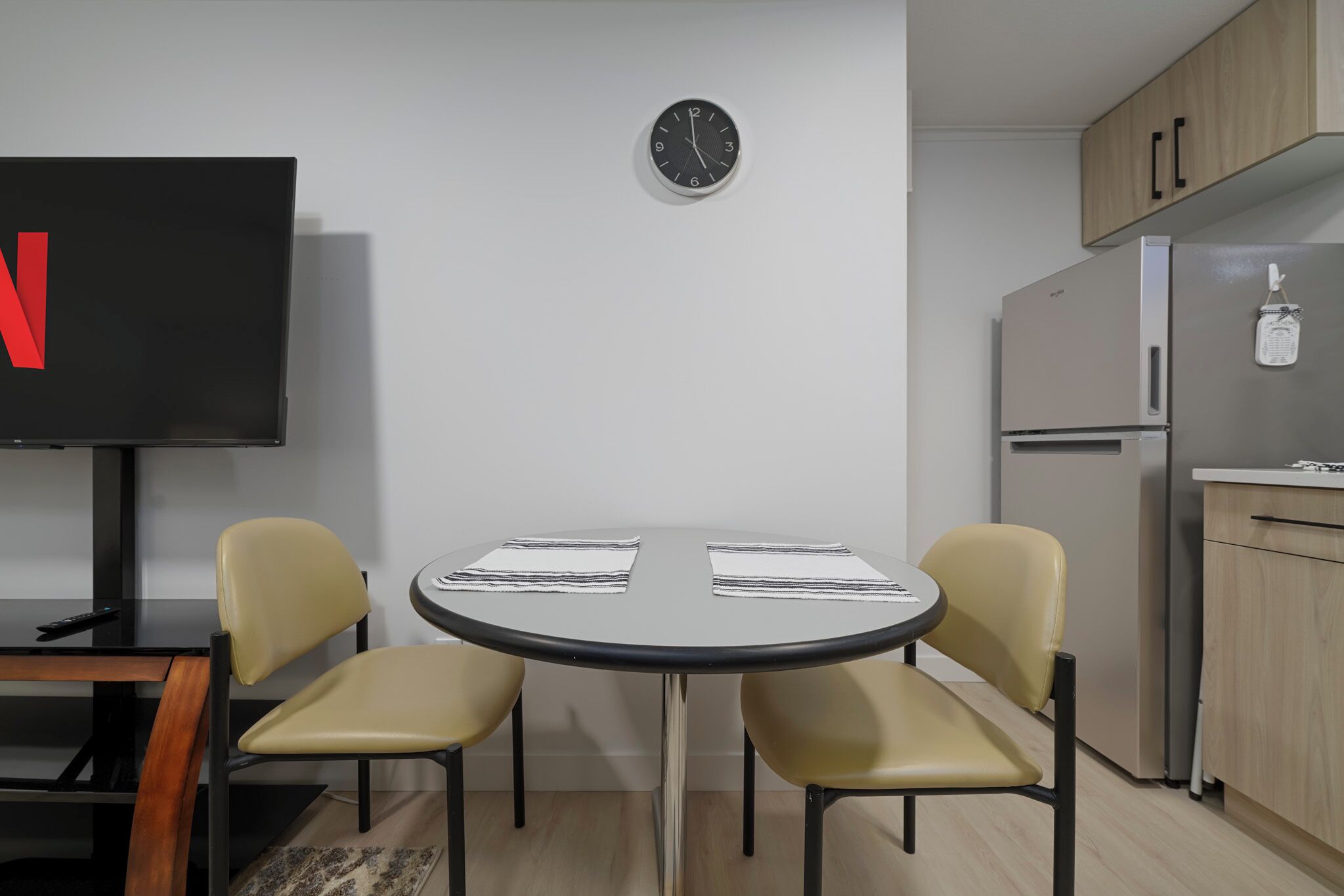 A modern kitchen with a round table, two chairs, a clock, a TV, and a refrigerator.