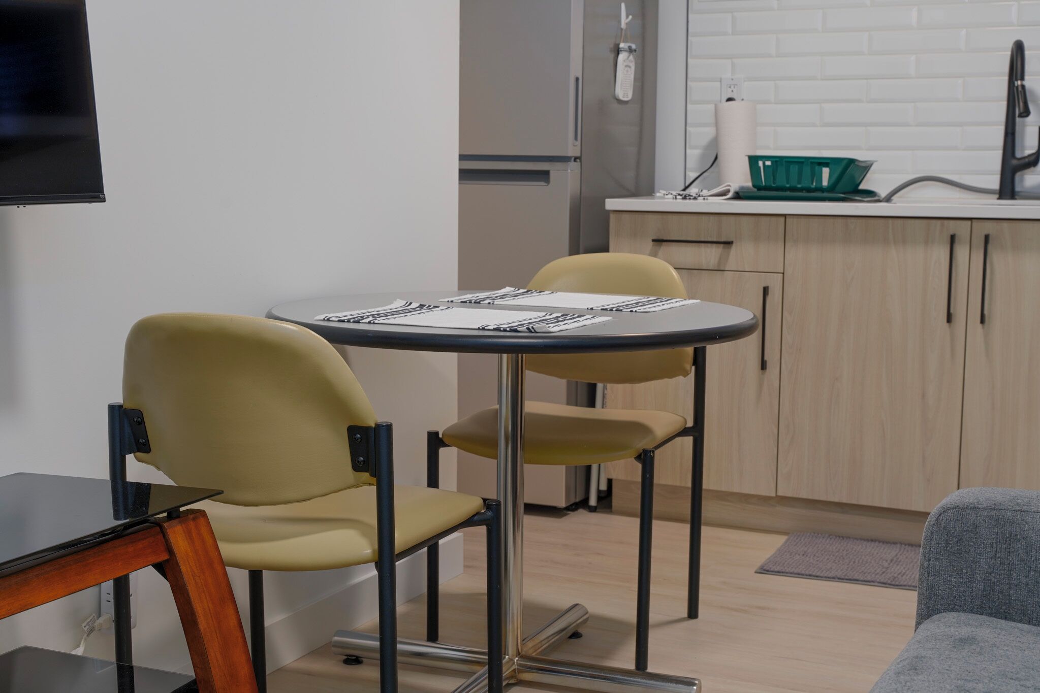 A compact kitchen with a dining table and chairs, a refrigerator, and a small countertop with a sink and a green basket.