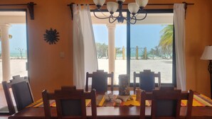 A dining area with a table set for four, large windows with curtains, a chandelier, and a view of a beach. - Ocean Front, Accessible, 3-bdrm Home!  (Celestún)