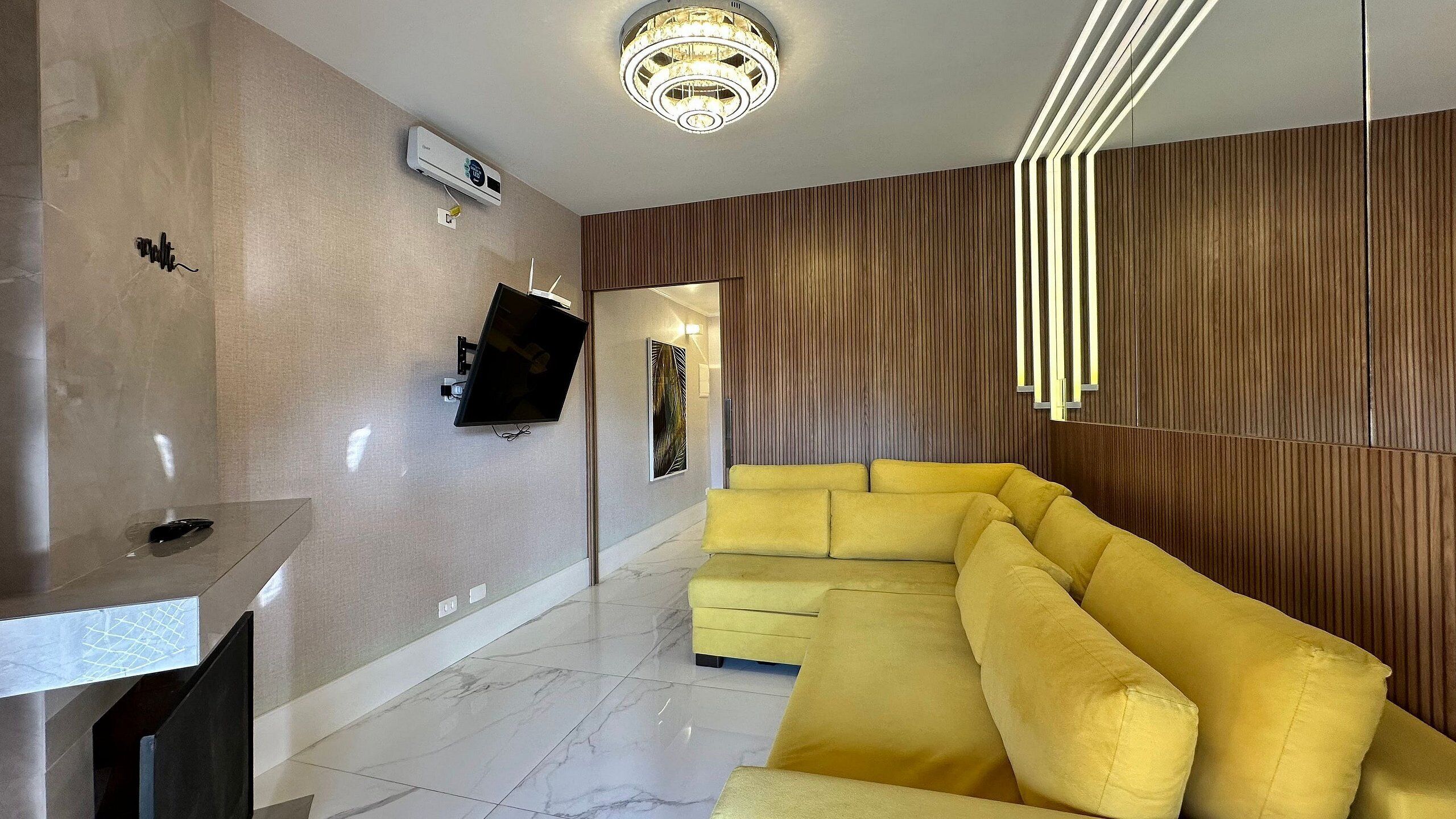 A modern living room with a yellow sectional sofa, a flat-screen TV mounted on the wall, a ceiling light fixture, and a marble-tiled floor.