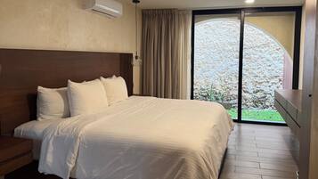 A hotel room with a large bed, a wooden headboard, a built-in air conditioning unit, a sliding glass door to a balcony, and a small wooden nightstand.