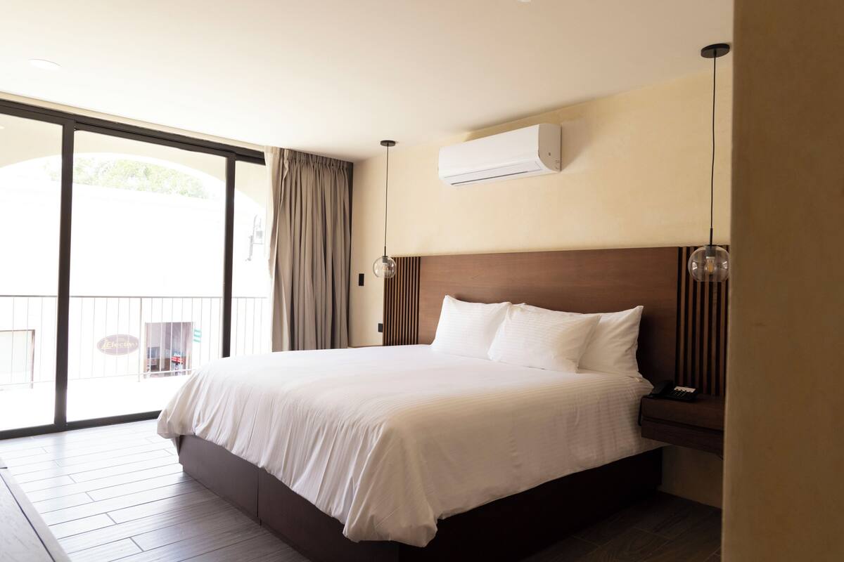 Ein Hotelzimmer mit einem großen Bett, einer Klimaanlage, einem hölzernen Kopfteil, einem Nachttisch mit Telefon und Blick ins Freie durch eine Schiebetür.