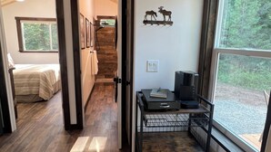A hallway with wooden flooring, a bed, a desk with a coffee maker, and a window with a view of trees. - Cozy cabin in charming forested area (Spirit Lake)