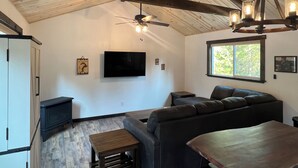 A spacious living area with a ceiling fan, a mounted TV, and a large window providing natural light. - Cozy cabin in charming forested area (Spirit Lake)