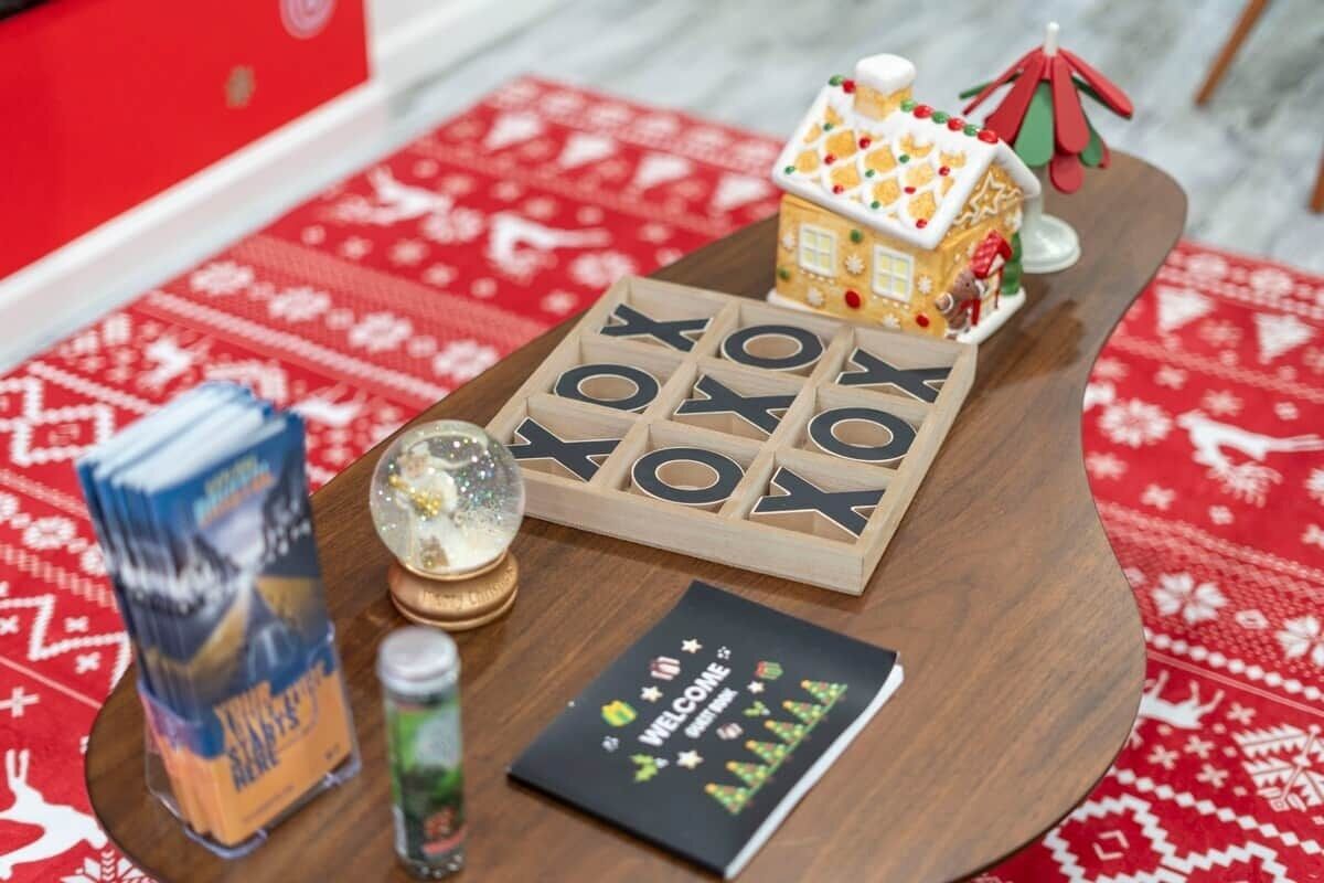 A wooden table with a wooden letter board spelling "WELCOME," a small gingerbread house, a snow globe, a box of milk, and a card with holiday decorations.