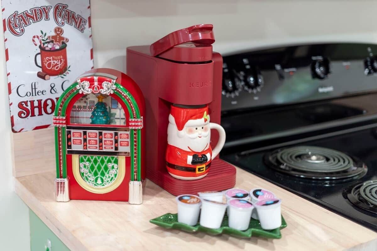 Una encimera de cocina con una cafetera roja, un pequeño modelo de tienda con temática navideña y tres tazas de café.