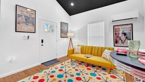 Un salon moderne avec un canapé jaune, un tapis aux motifs colorés et des affiches de journaux encadrées accrochées aux murs.