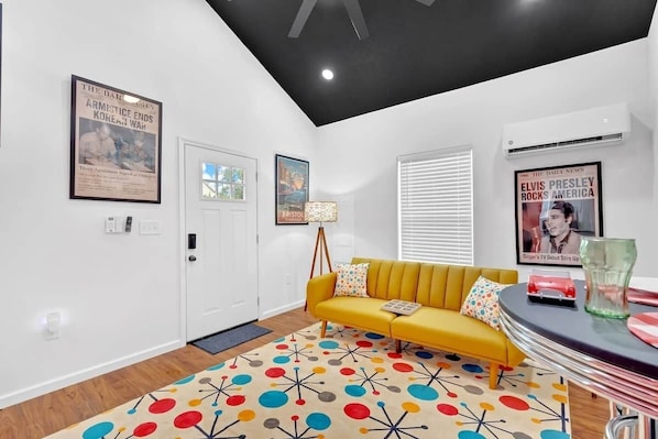 A modern living room with a yellow sofa, colorful patterned rug, and framed newspaper posters on the walls.