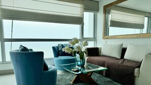 A modern living room with a glass coffee table, a blue armchair, a brown sofa, and a large window with blinds. - Exclusive apartment with sea view by Parceros Group (Cartagena de Indias)