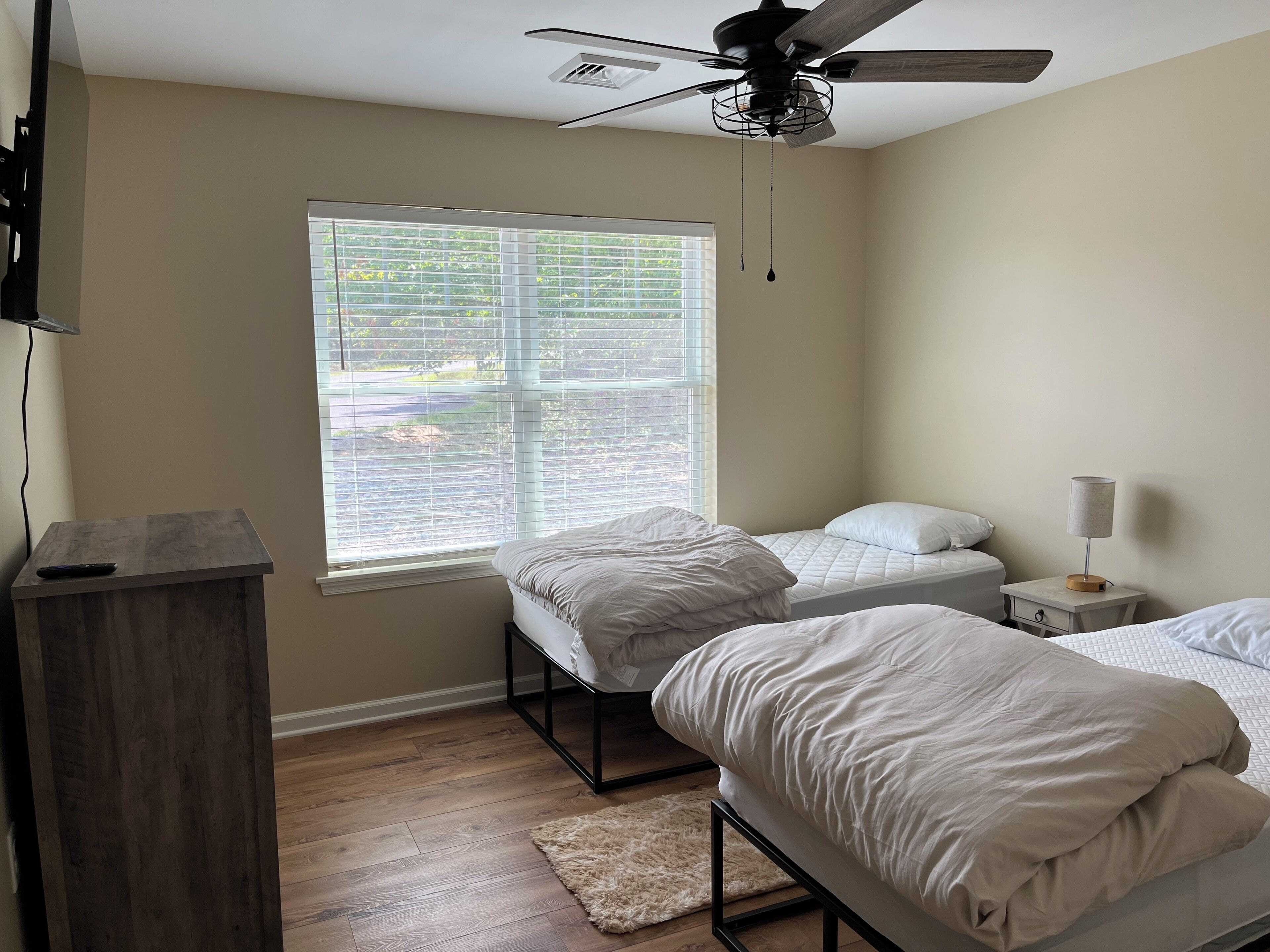 A bedroom with two beds, a ceiling fan, a window with blinds, a bedside table with a lamp, and a wooden dresser.