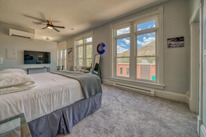 A bedroom with a large bed, a ceiling fan, a flat-screen TV, and a view of a building through large windows. - Downtown ~ Near River & Trails ~ Palace Hotel (Salida)