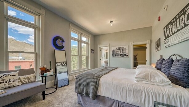A bedroom with a large bed, a bedside table, a mirror, and a window with a view of a building and mountains. - Downtown ~ Near River & Trails ~ Palace Hotel (Salida)