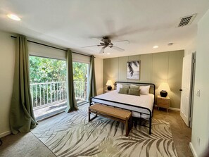 A bedroom with a bed, a ceiling fan, a nightstand with a lamp, a bench, and a view of greenery through a balcony door. - 316 Unit A - Welcome To Paradise! (Fort Pierce)