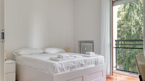 A white bed with towels, a radiator, and a view of trees through a balcony. - Iroko Living - Turro (Milano)
