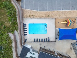 Vista aérea de uma área de piscina com piscina azul, espreguiçadeiras, área de recreação infantil e um espaço coberto para relaxar.