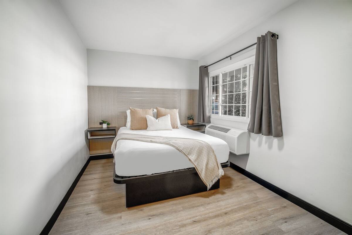 A modern hotel room with a large bed, a bedside table, a nightstand, a window with curtains, and a view of trees outside.