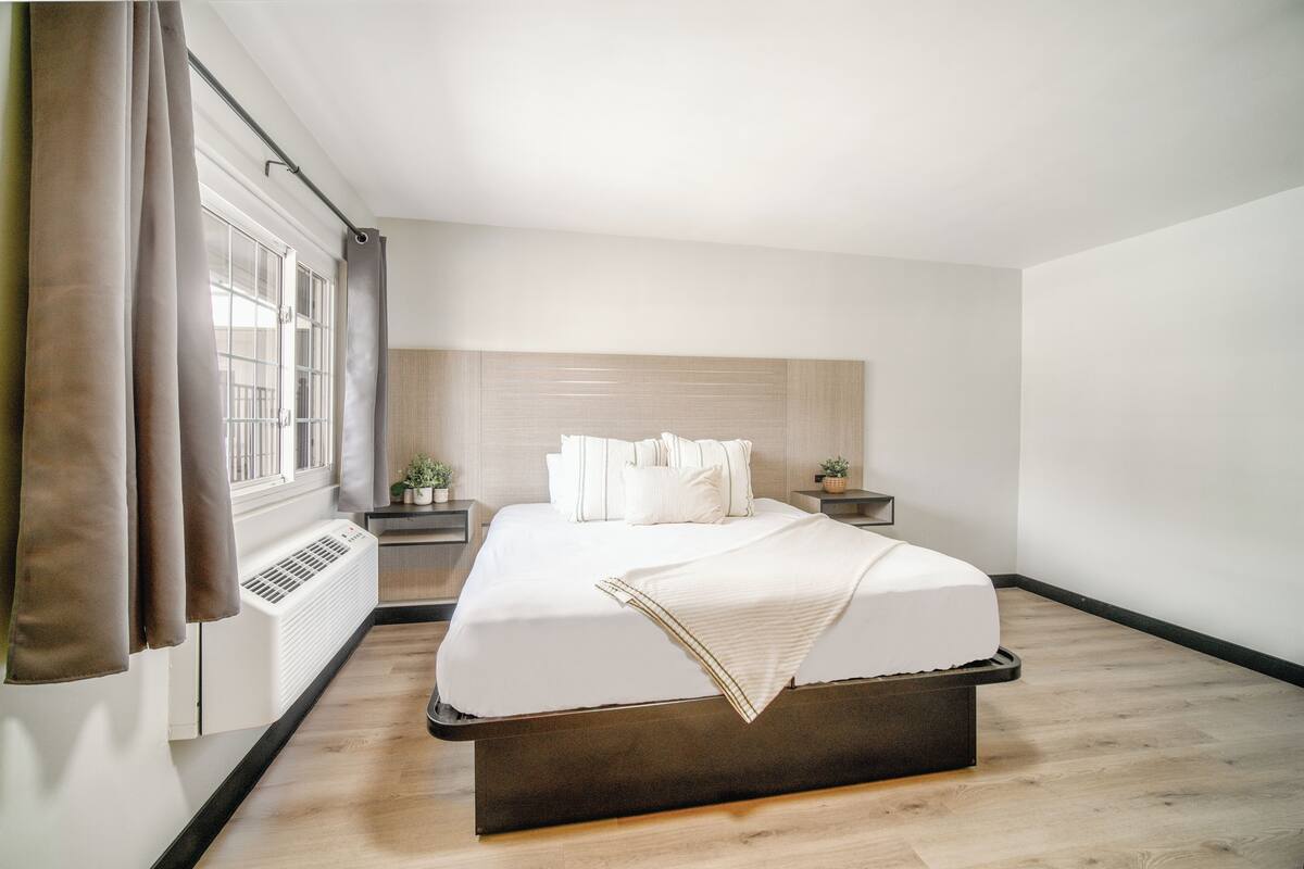 A modern bedroom with a large bed, a bedside table with a lamp, a window with curtains, and a wall-mounted heater.