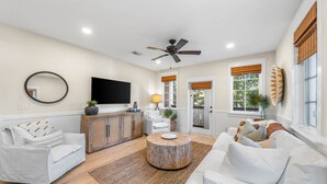 A spacious living room with a flat-screen TV, a ceiling fan, and a large window with blinds.