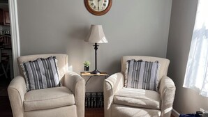 A living room with two armchairs, a small side table with a lamp, a clock on the wall, and a window with curtains. - The Red House  (Sodus Point)