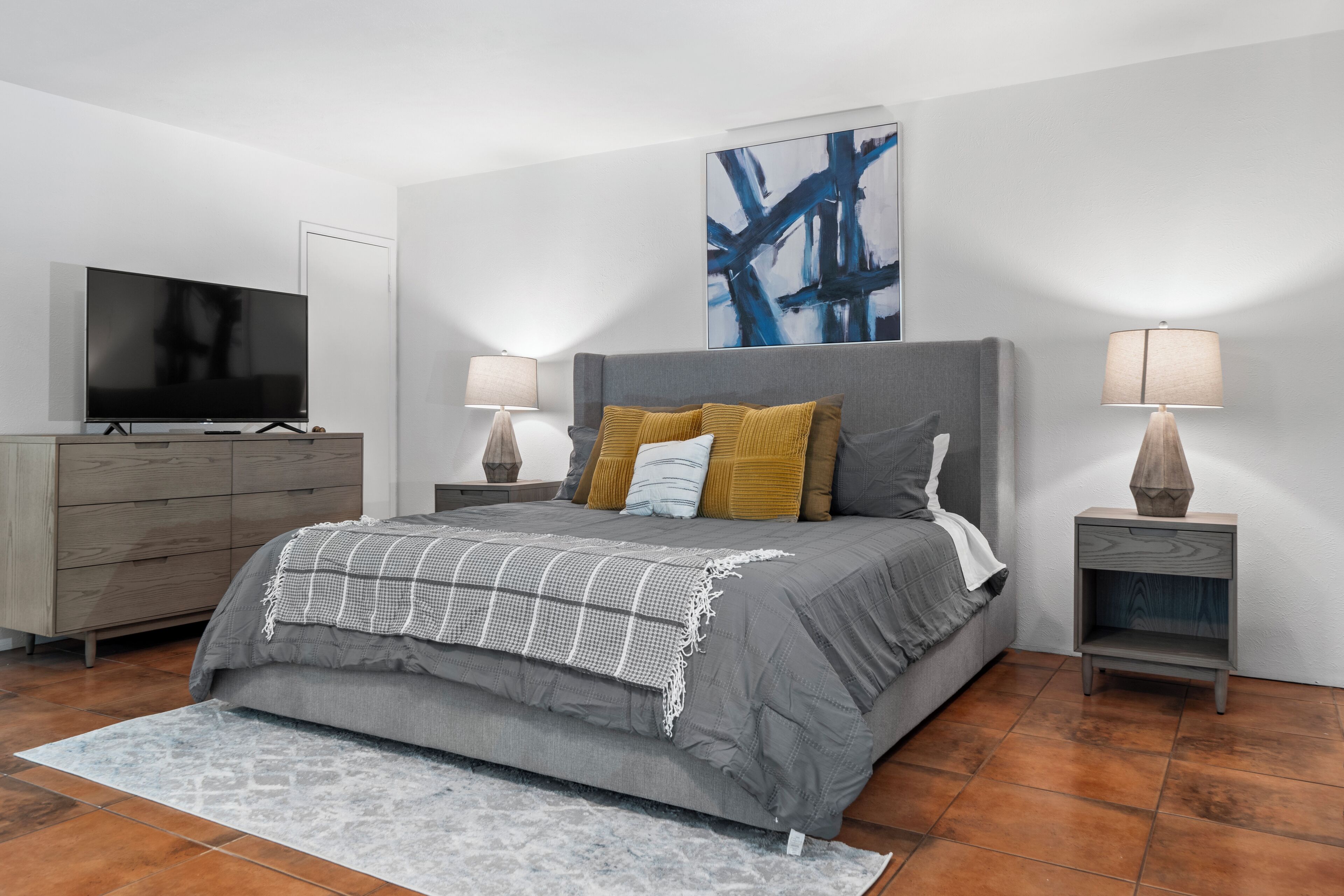 A modern bedroom with a grey bed, a flat-screen TV, two bedside tables with lamps, and a large abstract painting on the wall.