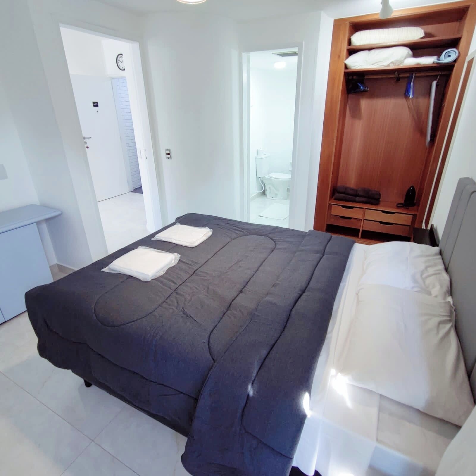 Une chambre à coucher avec un lit, une armoire en bois, une salle de bain visible à travers une porte ouverte et une petite table.