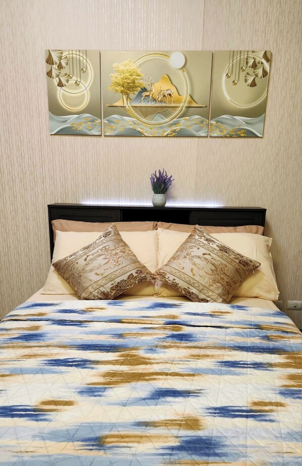 A bed with a patterned bedspread, two decorative pillows, a small potted plant, and a wall-mounted artwork above the bed.