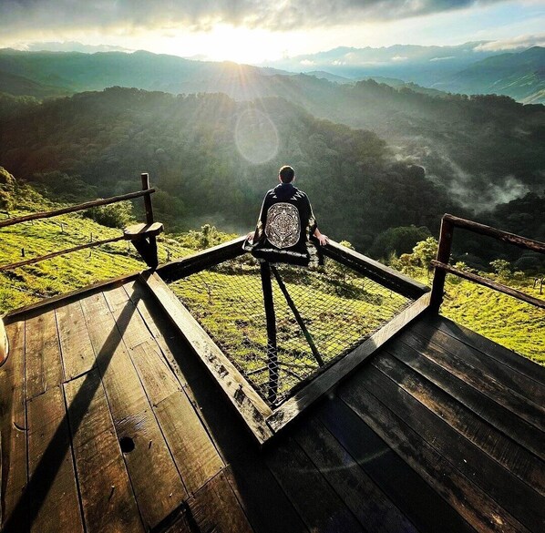 A person sitting on a wooden platform with a metal railing, overlooking a mountainous landscape with a sunrise or sunset. - Glamping Rustiko Salento  (Salento)