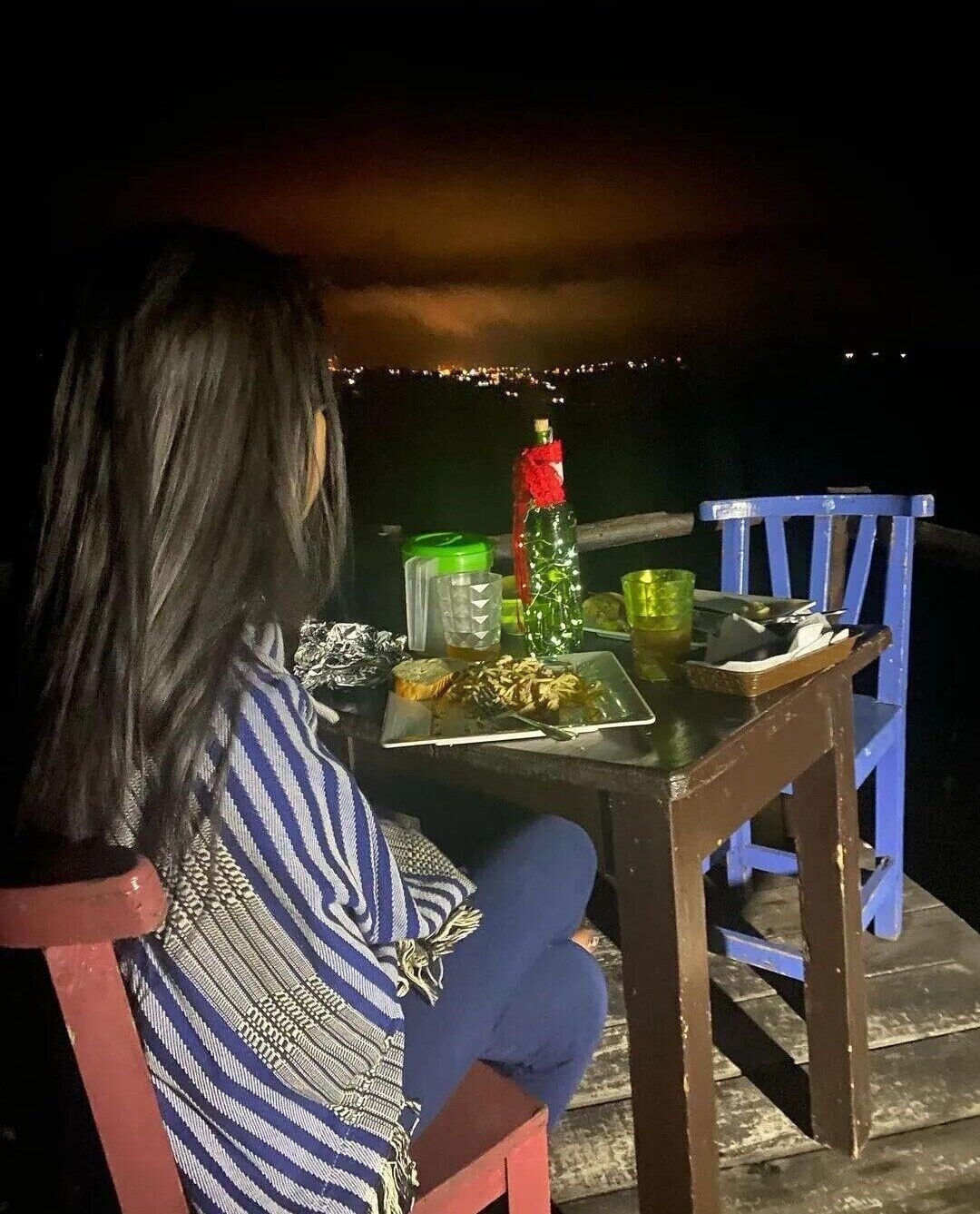 Una persona sentada a una mesa con un plato de comida, una botella y un vaso en una terraza de madera por la noche.