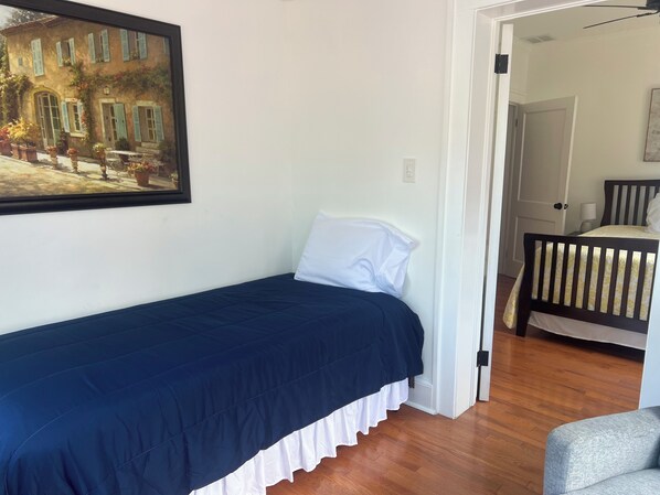 A small, neatly made bed with a dark blue bedspread and white pillow, a painting of a building with shutters on the wall, and a wooden floor. - Charming apartment with pool, free parking, walk to everything downtown. (St. Augustine)