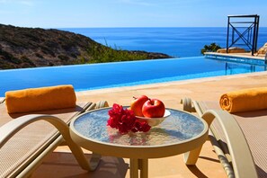 A round glass table with a bowl of fruit and flowers, two chairs with yellow cushions, and a view of a pool and ocean. - Luxury Villa Escape with Breathtaking Sea Views & Perks (Paphos)