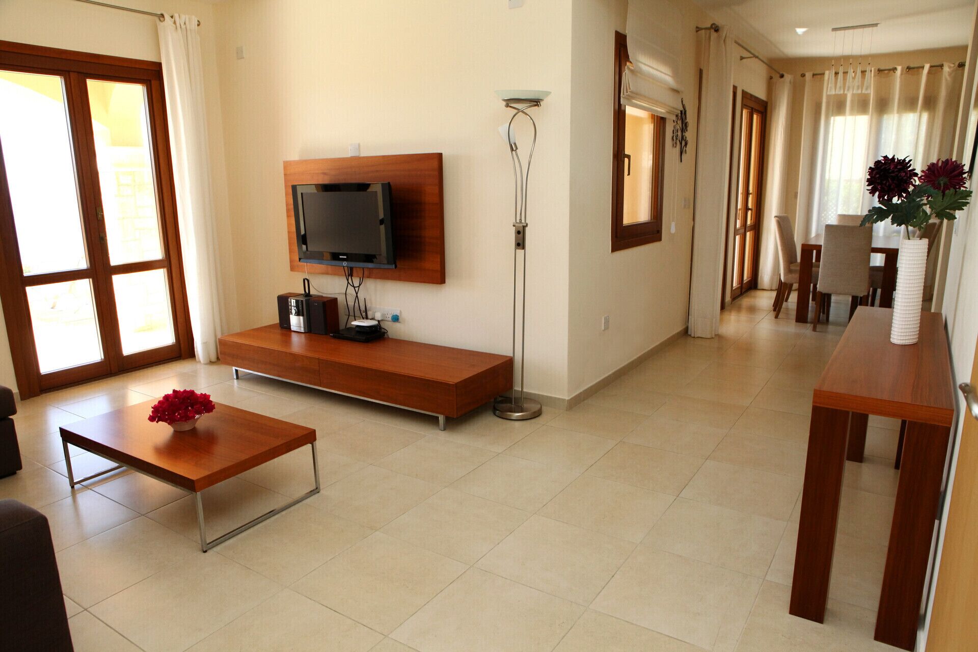 A modern living room with a TV mounted on a wooden wall, a floor lamp, a dining area with a table and chairs, and a small table with a vase of flowers.