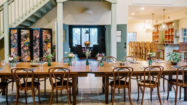 Un coin repas est aménagé avec une longue table en bois, dressée pour six personnes, et entourée de chaises en bois. Un escalier avec une rampe blanche est visible sur la gauche.