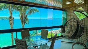 A modern dining area with a glass table and wicker chairs, overlooking a tropical beach and ocean. - Apt Luxo ao lado da Praia no Porto Real  (Mangaratiba)