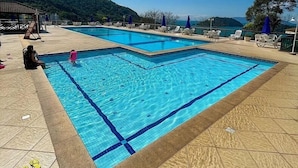 A swimming pool with clear blue water, surrounded by lounge chairs and umbrellas, set against a backdrop of hills and a clear sky. - Apt Luxo ao lado da Praia no Porto Real  (Mangaratiba)