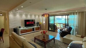 A modern living room with a large sofa, a coffee table, and a TV displaying 'NETFLIX'. There is a balcony with a view of the outdoors. - Apt Luxo ao lado da Praia no Porto Real  (Mangaratiba)