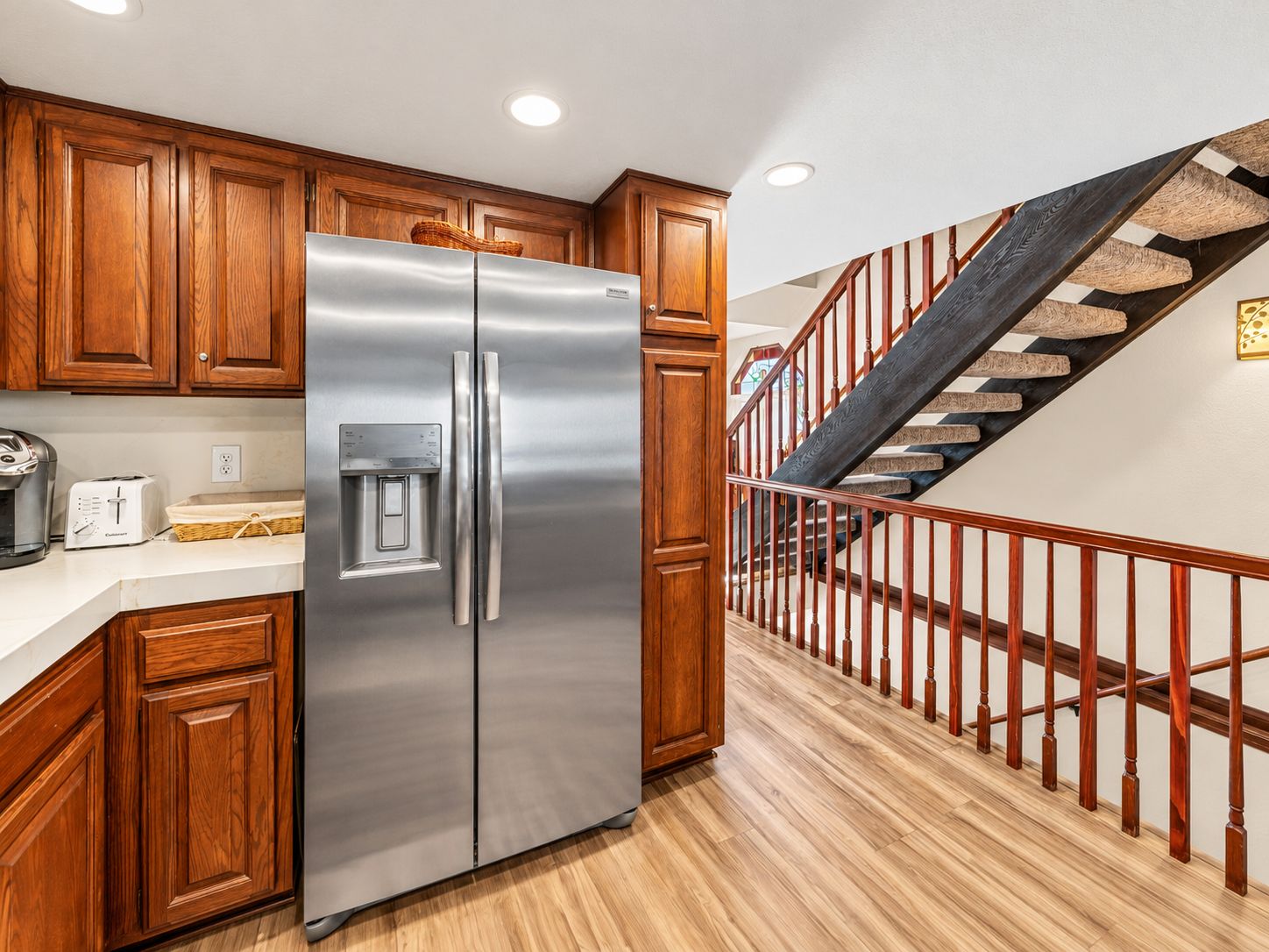 A kitchen with wooden cabinets, a stainless steel refrigerator, a water dispenser, and a wooden staircase with a railing.