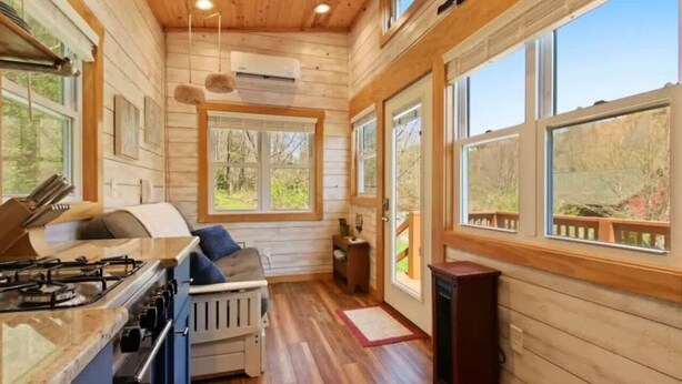 A cabin interior with a kitchen, a dining area with a table and chairs, a fireplace, and large windows offering views of the outdoors. - Charming Tiny Home! Discounts-Pet friendly (Todd)