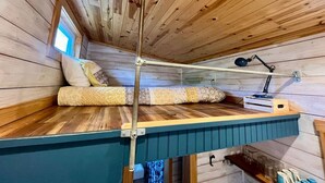A loft-style bedroom with a wooden ceiling, a bed with patterned bedding, a desk with a lamp, and a window. - Charming Tiny Home! Discounts-Pet friendly (Todd)