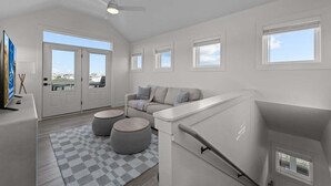 A spacious living room with a high ceiling, a ceiling fan, a sofa, two ottomans, a checkered rug, and a large window offering a view of the city. - Wright Off The Beach | 1020 ft to the beach | Private Pool (Kill Devil Hills)