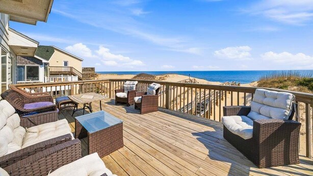 A wooden deck with wicker furniture, a table, and a view of the beach. - Fancy Free | Oceanfront | Hot Tub (Nags Head)