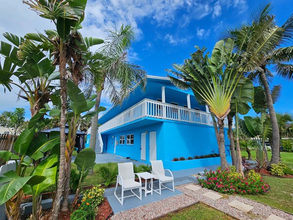 Una casa azul de dos pisos con un balcón, rodeada de plantas tropicales y un pequeño patio con sillas blancas.