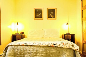 Een bed met een bloemenbedekking, twee bedlampen en twee ingelijste foto’s aan de muur.