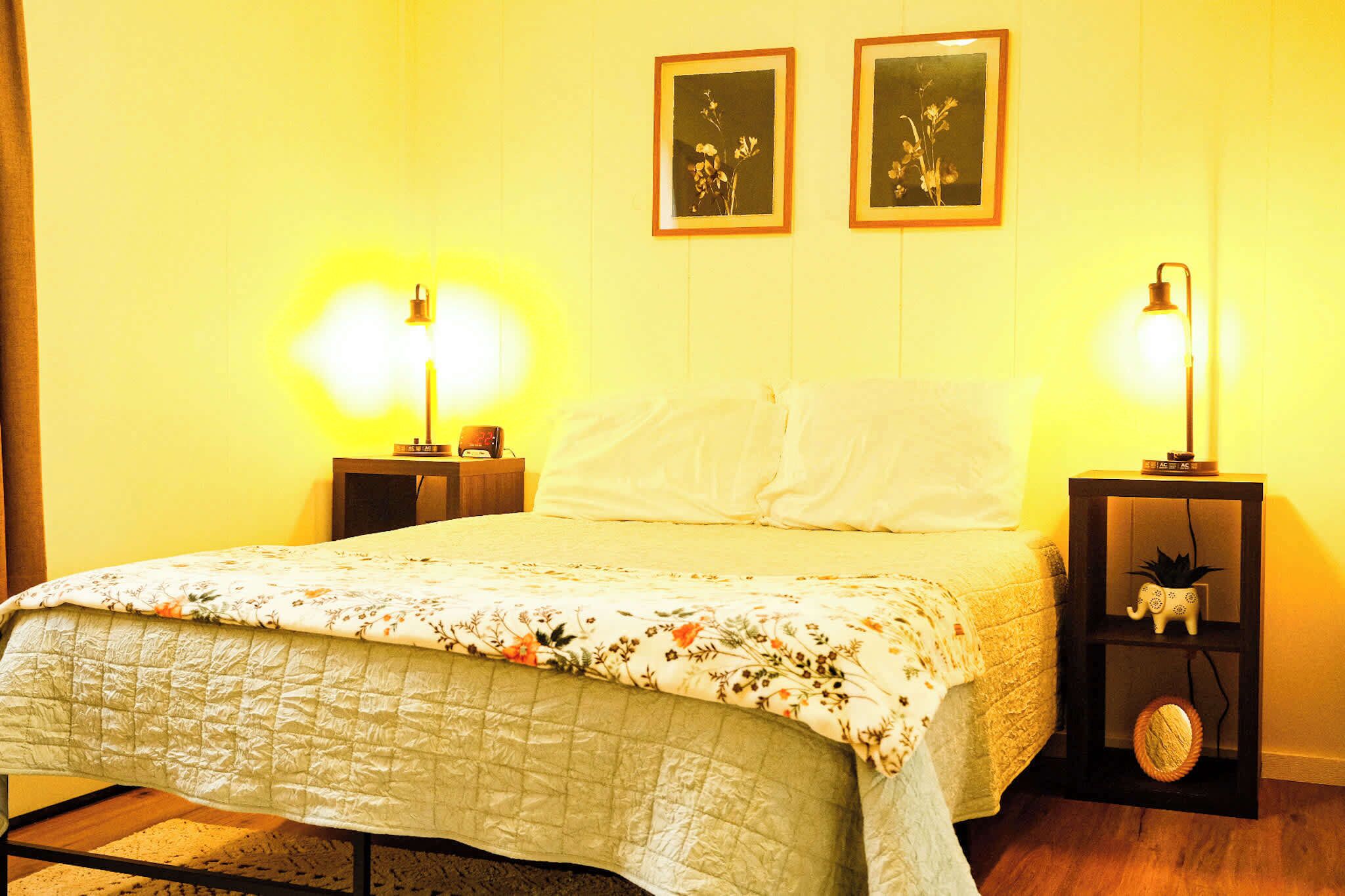 Ein Bett mit blumigem Überwurf, zwei Nachttische mit Lampen und zwei gerahmte Bilder an der Wand.